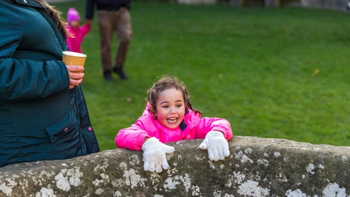 A family group outdoors with a close up of a smiling child leaning over an old stone wall in the foreground.